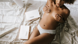 mujer acostada en la cama en topless, cubriendo su pecho, con cajas de regalo a su lado.